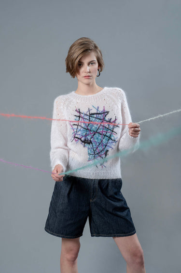 Woman wearing light pink mohair sweater and holding wool yarns going towards camera. The sweater has colourful embroidered lines forming an abstract shape in the center.